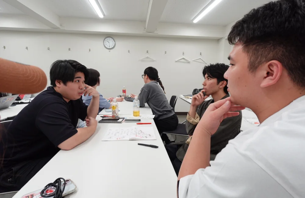 会議室のように長机を並べた部屋で、ACT-Rのメンバーが机を囲んで話している。机の上にはノートと図を書いた紙がある