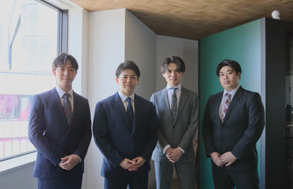 ACT-Rの男性4人が、窓と緑の壁のあるオフィスの一角に、スーツ姿で並んで笑顔で立っている