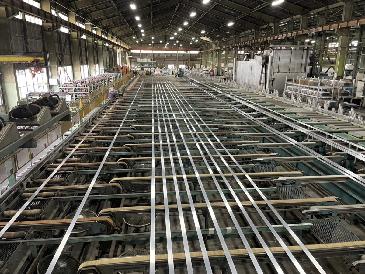 加藤軽金属工業の工場の中。長い台の上にアルミの細長い形材が何本も並び、奥で作業する人が小さく見える