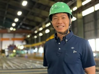 加藤軽金属工業 代表取締役 加藤大輝さん。緑のヘルメットをかぶり、紺のポロシャツ姿で笑顔を見せる。背後は工場の中