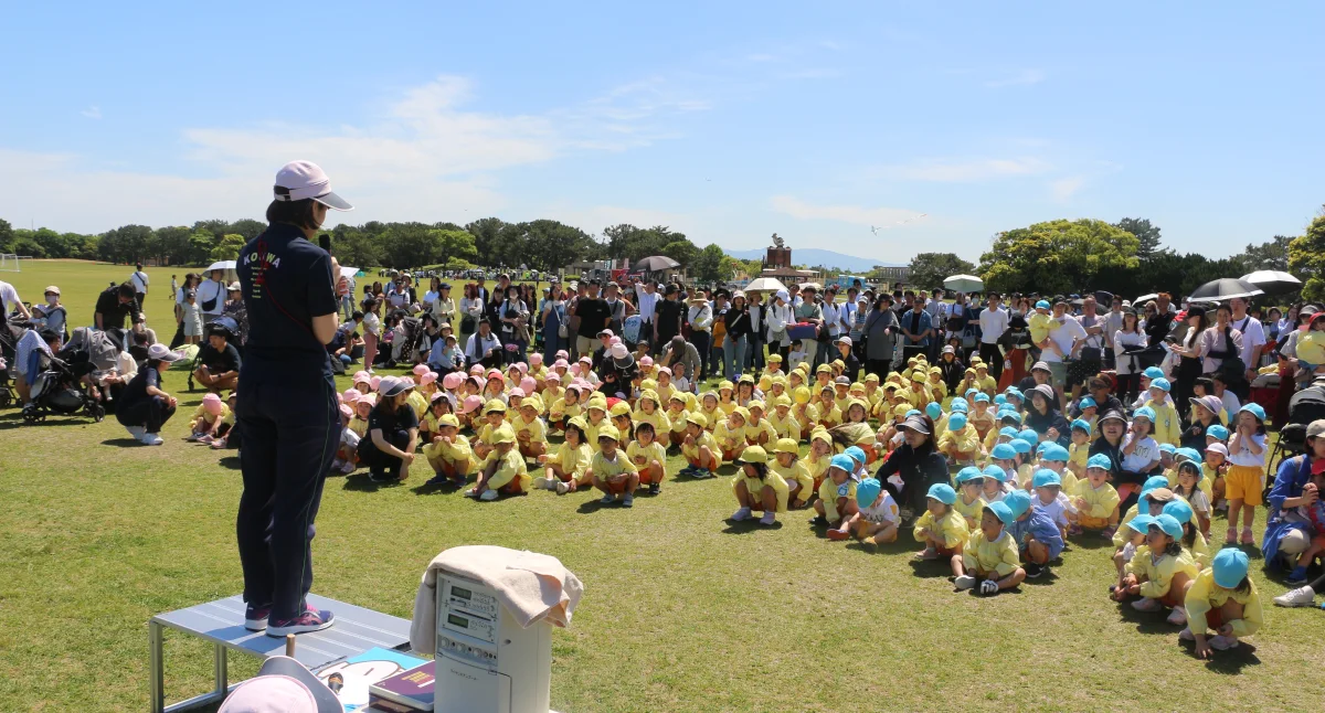 園庭の芝の上に、帽子をかぶった大勢の園児が座り、前で話す先生を見ている