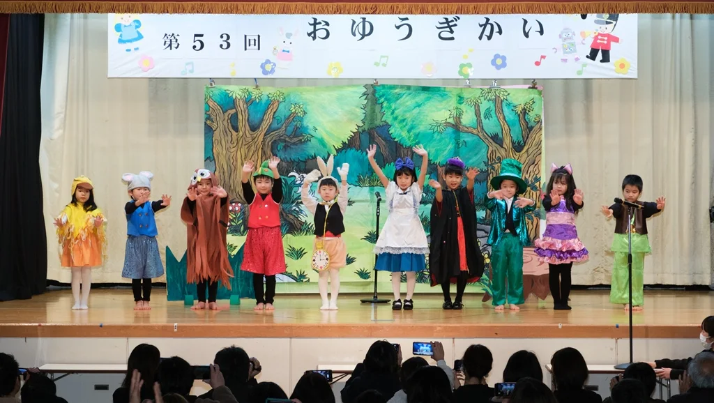 おゆうぎかいの舞台で、衣装を着た園児が横に並び、客席から見守られている