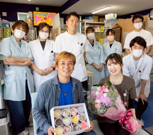薬局の中で、助川社長とスタッフ10名が集まって笑っている。社長は菓子箱を、隣のスタッフは花束を持っている